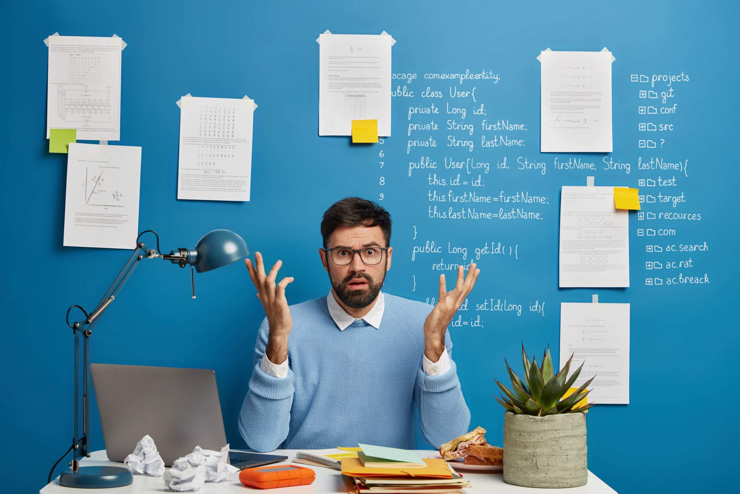 Confused bearded man raises hands with puzzlement, works freelance, uses laptop and papers, develops software, wears transparent glasses and blue jumper, searches useful information on website